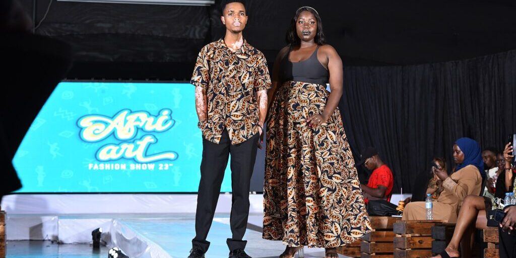 Two models in matching patterned African-print outfits stand on a runway at a fashion show, with seated audience nearby
