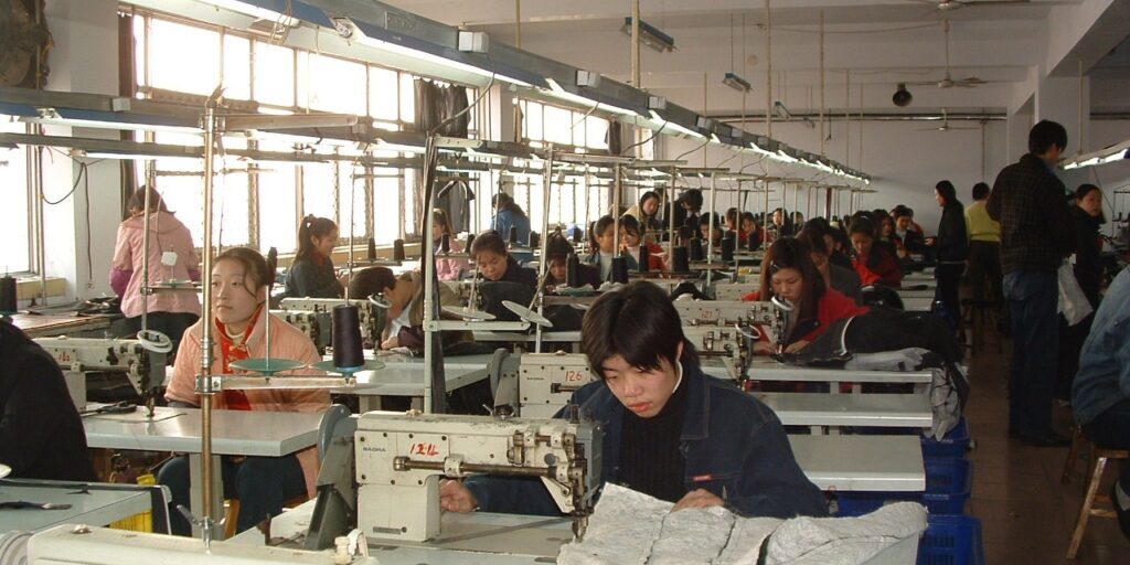 Rows of workers at sewing machines in a long, bright garment factory hall with fluorescent lights and ceiling fans