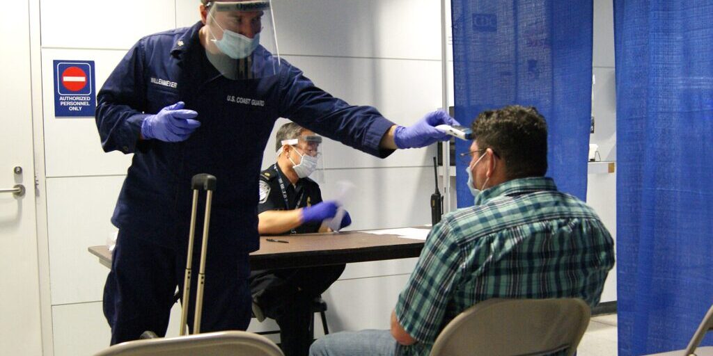 Masked, gloved Coast Guard corpsman holds a thermometer toward a seated passenger while an officer watches at a table