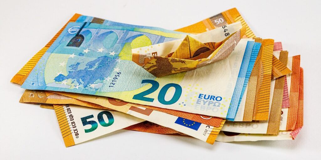 Euro banknotes in a loose pile, one folded into a small paper boat, on a white surface