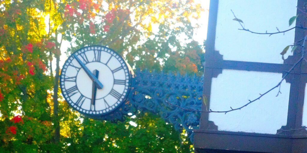 Round clock with Roman numerals on an ornate bracket beside a half-timbered wall, with autumn trees behind