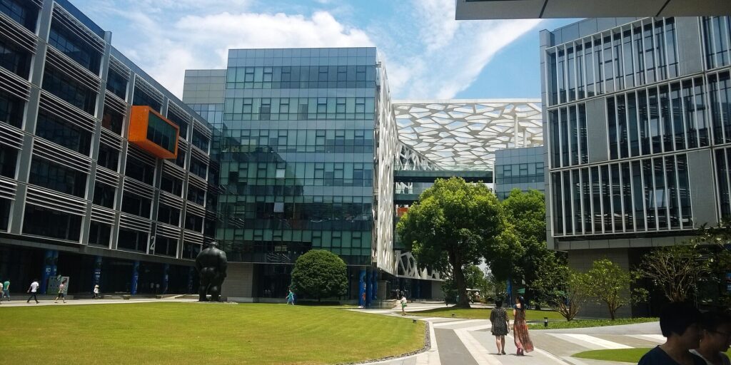Glass office buildings and a lawn on a campus, with a few people walking on the paved path under a blue sky