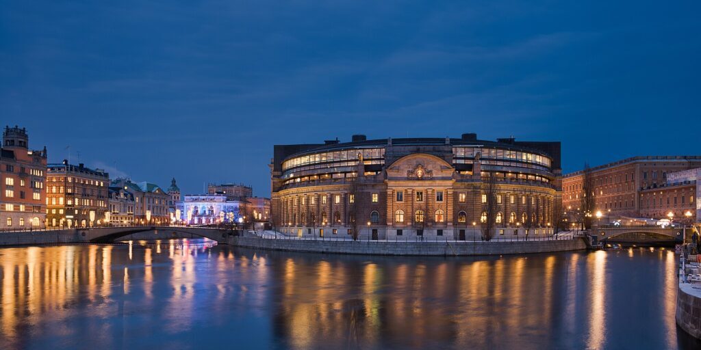 Riksdag House in Stockholm at dusk, its lit facade reflected in the water, with the Royal Opera in the background.