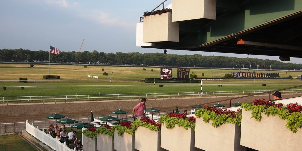 View from Belmont Park's grandstand overlooking the dirt track, infield, and scoreboard with an American flag flying.