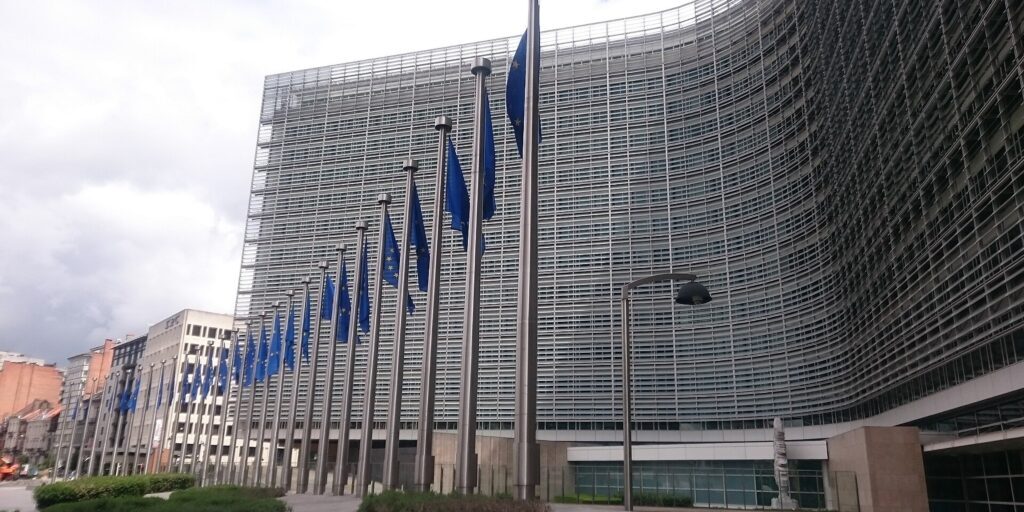 Row of EU flags on tall poles in front of the curved glass facade of the Berlaymont building under a cloudy sky