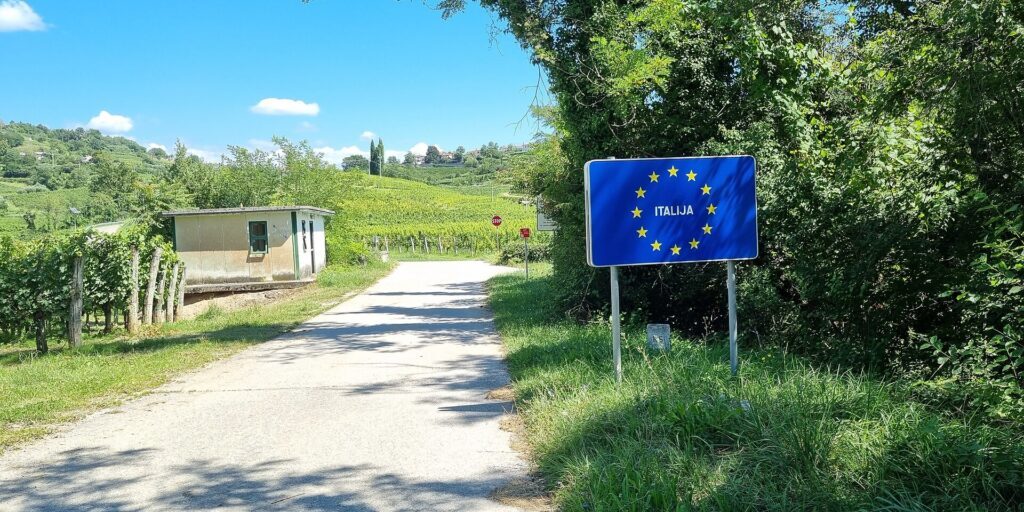 Rural road past a blue EU-star sign reading ITALIJA, with a small building and vineyards on a sunny day.