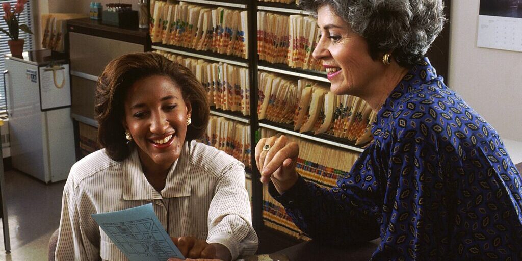 An older woman consults with a healthcare worker in a doctor's office with records in the background