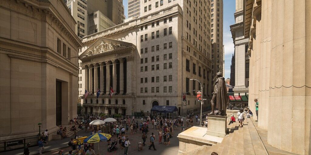 Crowded Wall Street intersection with pedestrians and a street vendor's umbrella, beside a columned stock exchange building