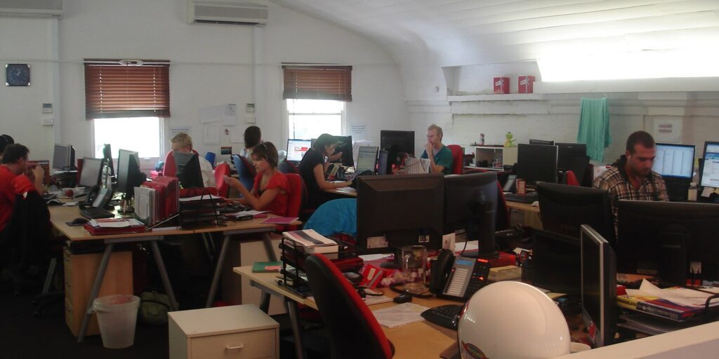 Workers at desks in a bright open-plan office with computer monitors and red chairs.