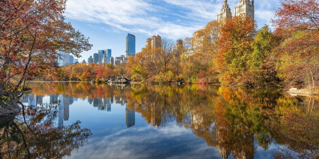 Autumn trees with orange and green leaves around a calm lake, reflecting a city skyline and a tall stone tower under blue sky