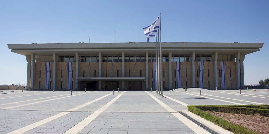 Front view of the Knesset building in Jerusalem under a clear blue sky, with Israeli flags on poles in the foreground