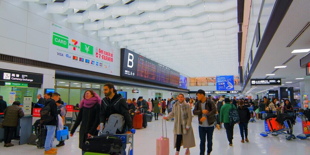 Travellers with luggage walk through a bright airport arrivals hall with currency exchange signs and a row of check-in counters.