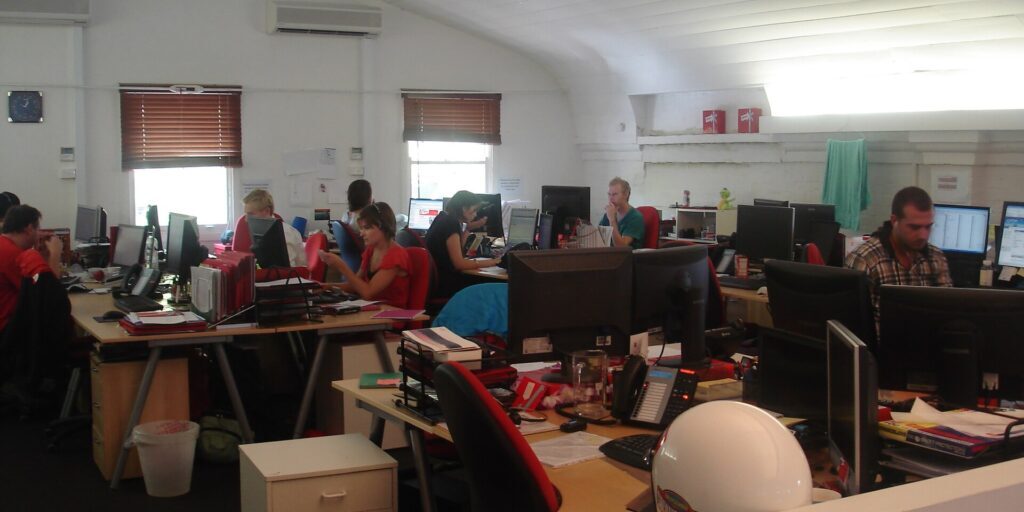 Office workers at computer desks in an open-plan office with red chairs, monitors and telephones.