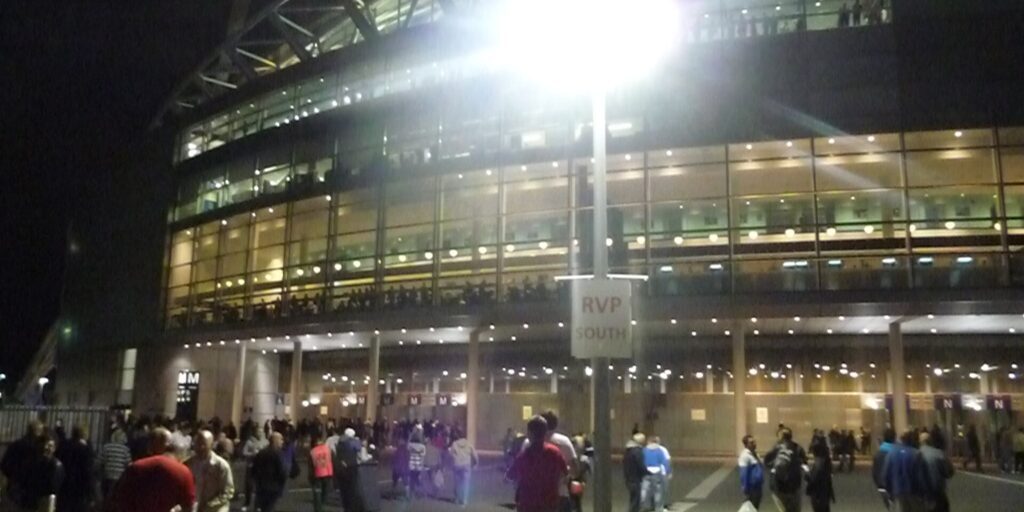 Crowds of people gather outside a lit stadium entrance at night, with a floodlight and an RVP South sign.