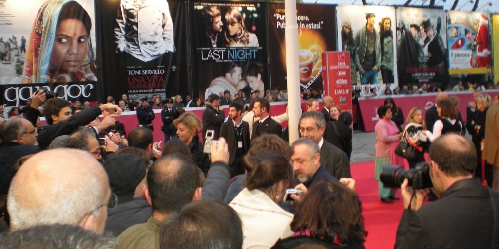 Crowd of photographers and onlookers beside a red carpet, with large film posters on the wall behind