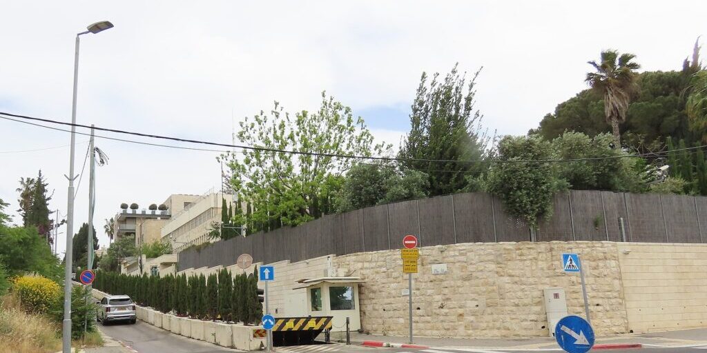 Stone-walled exterior of a building behind a security wall, with a guard booth and a road in the foreground