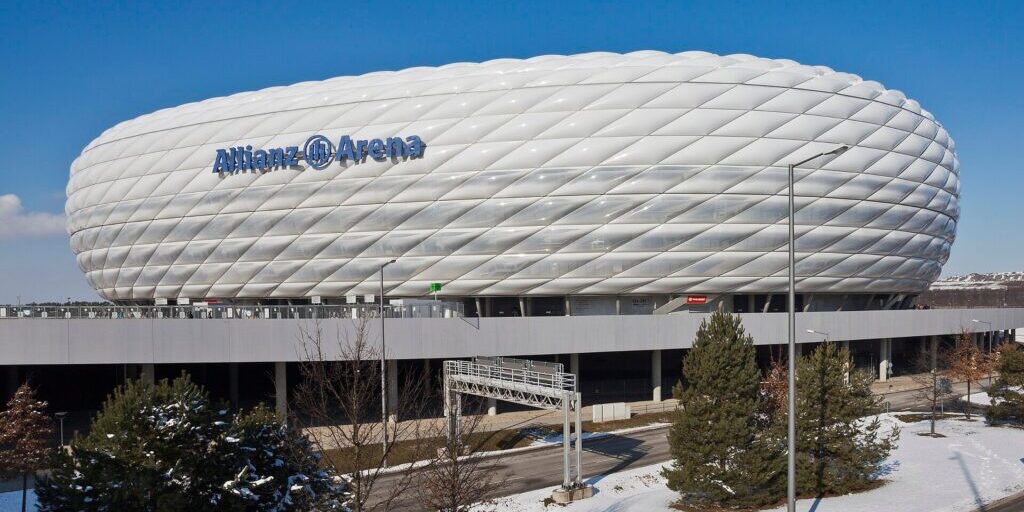 Exterior of the Allianz Arena stadium in Munich under a clear blue sky, with snow on the ground and trees in the foreground.