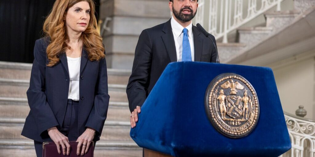A man speaks at a blue podium bearing a city seal while a woman holding a folder stands beside him on a staircase