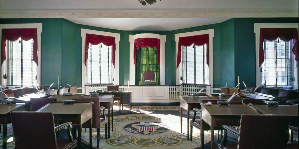 Historic Senate chamber with green walls, desks, and draped windows at Congress Hall