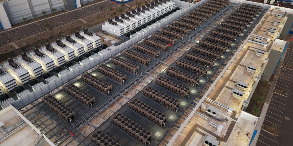 Aerial view of a data center roof with rows of cooling equipment and lights, beside a parking lot and construction site