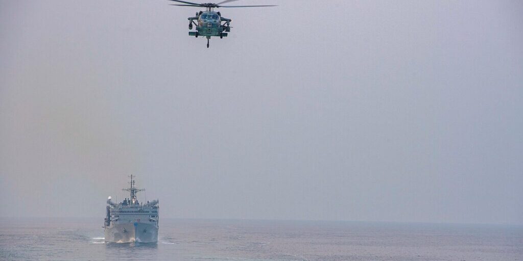 Grey military helicopter flies above a navy supply ship on hazy open sea, seen from a flight deck edge.