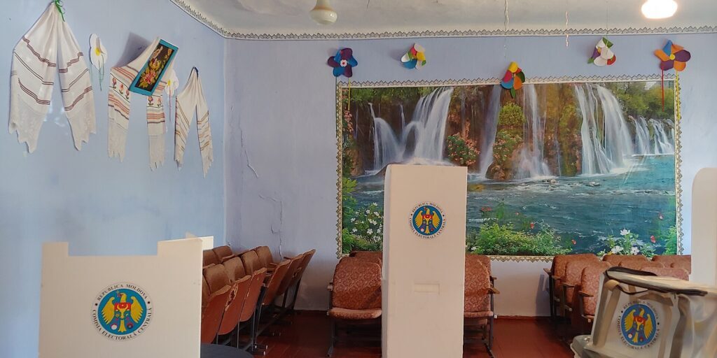 Empty polling station room with cardboard voting booths, a large waterfall wall painting, and rows of chairs along the wall.