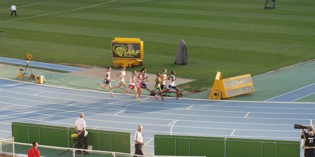 Runners competing in a middle-distance race on a blue track in a stadium, with spectators and officials along the rail.